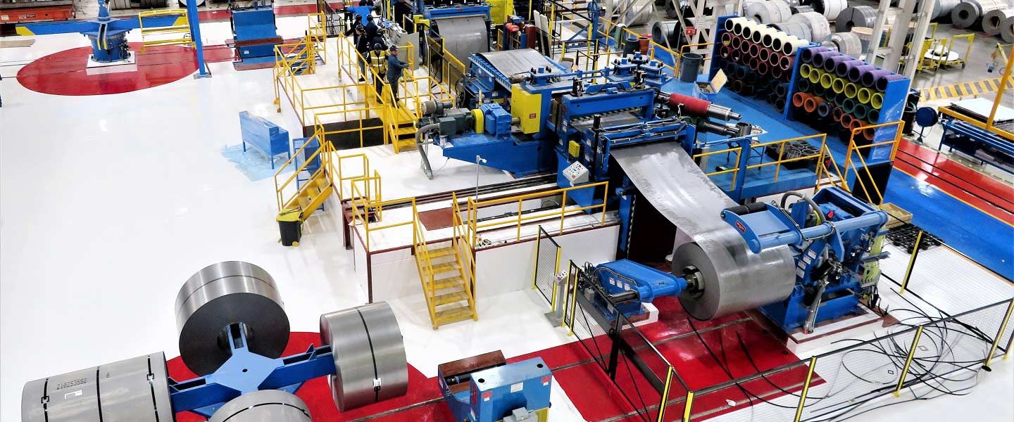 Large coil processing line in factory with blue machinery and yellow safety rails handling rolled sheet metal.